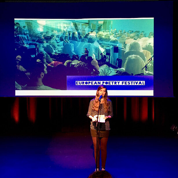 A picture showing a woman standing at a microphone in front of a screen that says "European Poetry Festival"